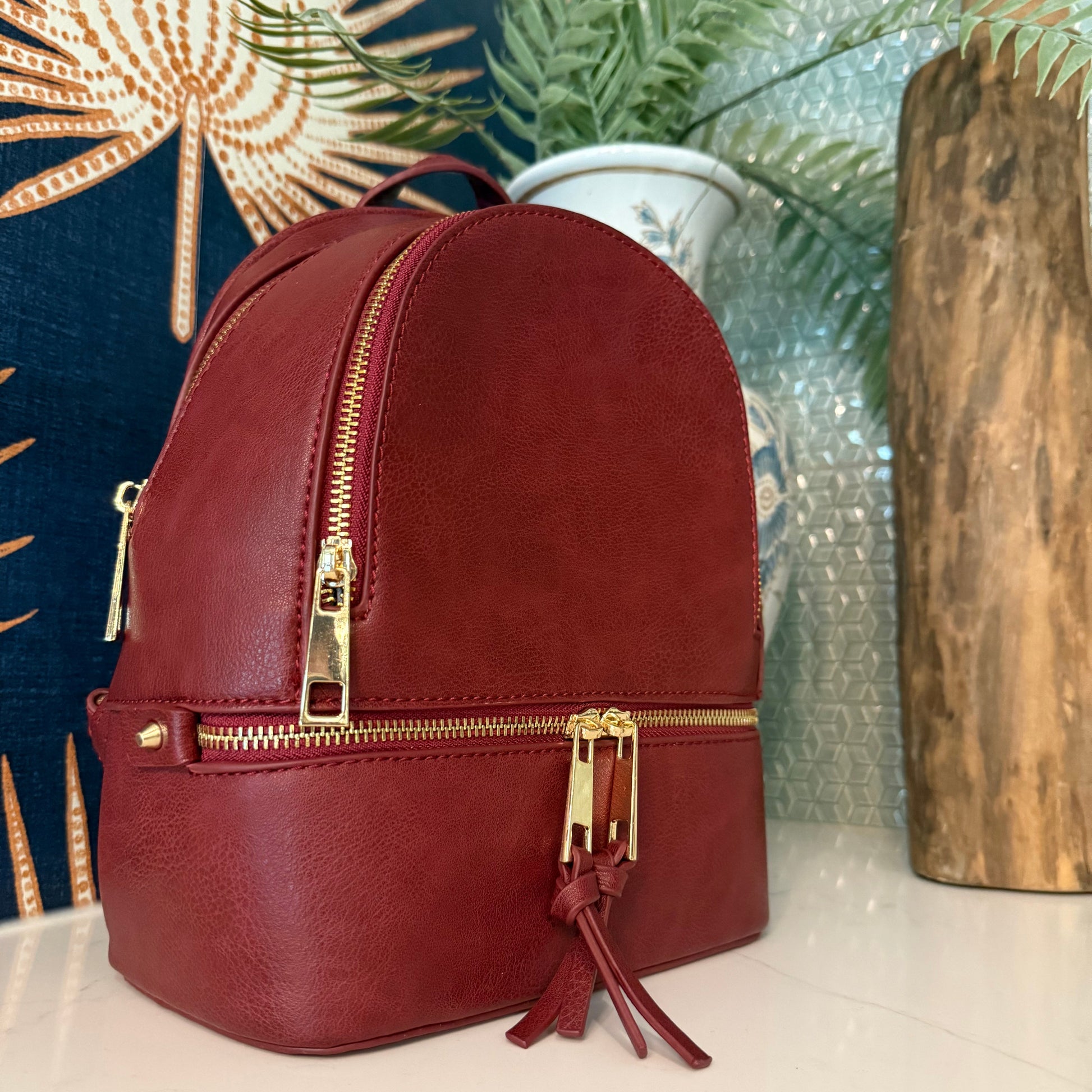 Burgandy Backpack Small with gold zipper on a textured surface with a plant in the background