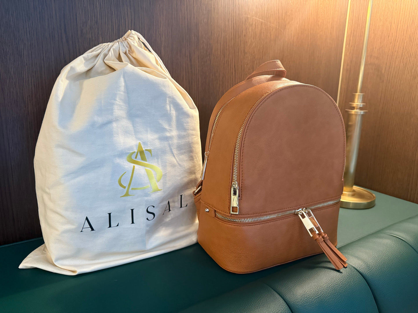 Tan leather backpack next to a white drawstring bag with 'ALISAL' branding on a green surface.